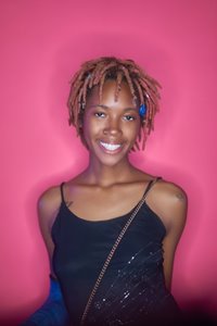 A brown-skinned 21-year-old black woman smiling. Her hair is styled in pale pink locs with blue seashell hair jewelry. She's wearing a black dress, but you can see two tattoos: one that reads "222" on her right shoulder and a tulip on her left shoulder. The background is bright pink and shows Imani's shadow.