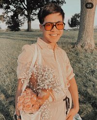 A happy young man posing in a grass field carrying a tote bag filled with flowers and a baguette. He is wearing red-tinted sunglasses, a peach-colored shirt, and beige-colored shorts.