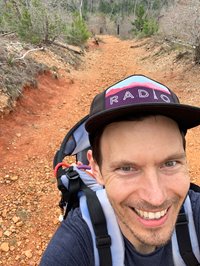 Person with large smile in lower left corner of the photo has light white complexion, short brown hair, and stubble. They are wearing a dark blue t-shirt and black baseball cap with a purple and magenta  patch that reads "Radio." They are also wearing a light purple backpack with a child on their back. In the background is a rocky trail with orange rocks and dirt. Pine trees can be seen off in the distance.