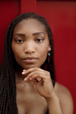 A warm-brown skinned femme in front of a vivid red background looks directly at the camera, hand resting against chin. Naiya has long dark brown box braids, a black top, and vibrant turquoise and copper earrings.