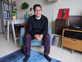 Aimee sits in her home with a big, welcoming smile on her face. She is wearing a navy sweater, under which a white collar frames her face, purple pants, and navy socks. Behind her are books, records, a lamp, and some houseplants.