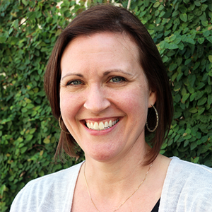 Sarah Rinner headshot. Sarah looks into the camera with a big, warm smile. She has light skin and shoulder-length straight brown hair, and wears hoop earrings, a thin chain around her neck, and a white top with a black top visible underneath it. Behind her is green foliage.