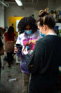 One brown skinned youth with black curly hair, and one white woman with brown and blonde hair in a bun are holding containers of paint, mixing colors. They are in a dimly lit art studio environment with concrete floors.