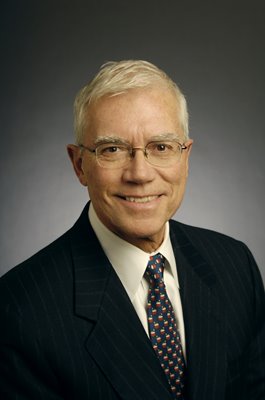 Terry is smiling into the camera, wearing glasses with thin wire frames, a dark suit jacket, a white shirt, and a navy blue tie with a red pattern on it. He has light skin and white hair. The background is a black and grey gradient.