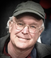 A closeup of Jeff with a dark background. He has light skin and short, white hair, and wears a dark cap, wire-rimmed glasses, and a dark jacket with a shirt collar visible underneath.