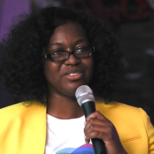 Photo of Quanice G. Floyd. She is smiling while holding a microphone in her hand, wearing black rectangular-framed glasses, a bright yellow blazer, and a white t-shirt with a colorful design on it. She has brown skin and curly black hair. The background is black and dark purple.