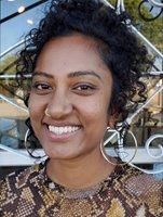 A closeup of Raji smiling brightly into the camera. She has medium brown skin, loose black curls falling around her face, and is wearing a snake-skin patterned top, large gold hoop earrings, and an ornate gold nose ring.