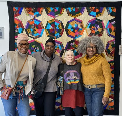 three black women and one white woman pose in front of a multi-colored quilt