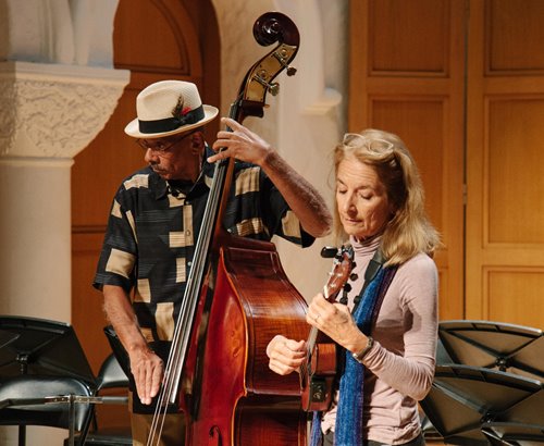 Two older adults performing with strong instruments. The person on the left has light brown skin and is wearing a hat and a patterned shirt. The person on the right has light skin and long, blonde hair, and is wearing a light purple turtleneck. Photo courtesy of Pasadena Conservatory of Music