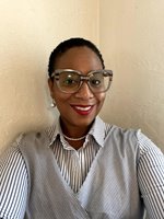 A selfie of Patricia in front of cream-colored walls. She has brown skin and dark brown hair cut close. She wears large, thick-framed grey glasses, red lipstick, a white dangling earring, a pearl necklace, a button-down shirt with white and dark blue vertical stripes, and a grey wrap vest.
