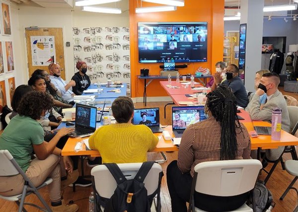 Board and staff gathered around tables in a colorful room, with a large screen on the wall with virtual participants' Zoom squares