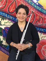 Martha leans against a wall adorned with a colorful mural, smiling and resting a flute on her shoulder. She has light olive toned skin and short, dark curly hair, and wears a necklace with a large silver pendant, silver bracelets, a black top, and dark pants.