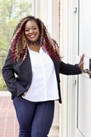 Calida stands with her hand resting on a white door, with a patio and trees in the background. She has long brown, blonde, and red curly hair, brown skin, and is wearing a white button-down shirt with a black blazer and dark denim pants.