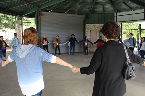 A group of people standing in a circle with their hands joined, outdoors, underneath a structure with a green domed ceiling.