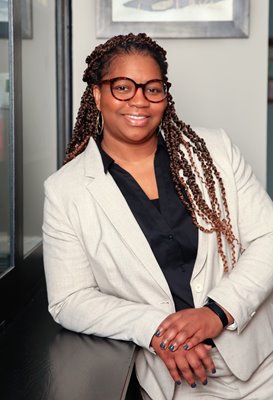 a brown woman with glasses and shoulder-length braids poses wearing a tan suit and black shirt