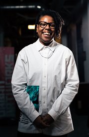 A headshot of a brown skinned nonbinary person with dreadlocks, looking into the camera with a big smile. They are wearing a white collared shirt, silver necklace and black rimmed glasses. Their dreadlocks are to the side of their face.