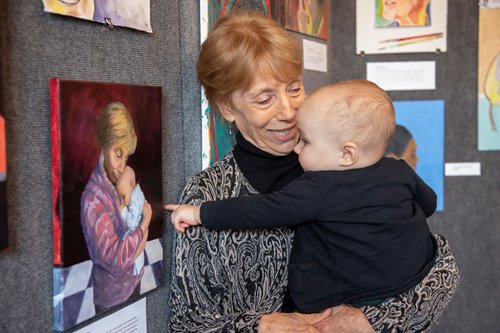 An older adult smiles at a baby they are holding. The baby is pointing at a painting on the wall, which is of the same older adult holding the same baby.