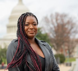 Portrait photo of Quanice G. Floyd. She is wearing a black jacket and has red and black braided hair. The Capitol building can be seen behind her.