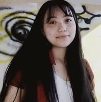 Winter stands with a small, closed-lip smile in front of artwork with yellow and black lines. They have long, dark, straight hair with soft bangs, and light skin with freckles. She is wearing a red, short sleeved shirt that is open to reveal a white top underneath.