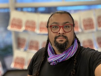 Headshot of a light brown Mexican-American male with pulled back hair with two braids down to the chest. William has a goatee and long side burns. He is wearing two silver small hoop earrings and cherry red glasses and he has a piercing on his right nostril and bottom left lip. He is wearing a black t-shirt and a multicolored stripped “reboso” or long woven scarf wrapped around his neck. The background is blurred and shows multiple screen printed posters on white paper that high light a quote by Grace Lee Boggs that says, “The only way to survive is by taking care of one another.”
