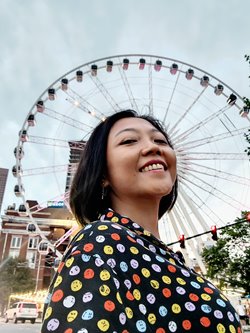 Smiling, olive-complexioned Southeast Asian woman with dark-brown hair in a short bob cut, wearing a black shirt dotted with colorful smiley faces, in front of a large ferris wheel that creates an illusion of a halo around her head.