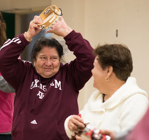 An older adult in a maroon-colored sweatshirt holds a tambourine above their head and smiles at another older adult, who is smiling back at them and facing away from the camera.