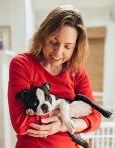 Kate is wearing a red shirt  in a room with white walls and a white dog pen behind her. She is holding Grover, a black and white puppy. She has frizzy light brown hair, and is smiling at the dog.