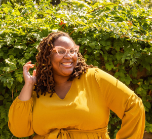 Precious is standing against a leafy backdrop wearing a mustard yellow top and light orange triangle shaped glasses