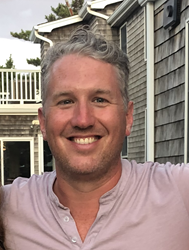 Adam is outdoors, with a wide smile and a light purple shirt. He has short, wavy grey hair and light skin, and there are houses in the background.