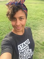 Ashley is standing in a grassy field, wearing a purple and pink scarf tied around their head, large silver hoop earrings, and a t-shirt that says "Stand with Black women". They have light brown skin and loose curls, and smiling with their mouth closed while holding up the camera.