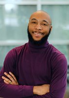 Jordan folds his arms and smiles gently, with a light blue glass window behind him. He has caramel colored skin, a shaved head, and a dark, full beard. He wears wire-rimmed glasses and a deep purple turtleneck.