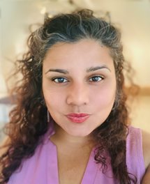 Close up of a smiling, tan female of South Asian descent with curly brown hair with grey streaks wearing a pink sleeveless top.