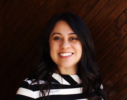 A person with light brown skin and long black hair, standing in front of a deep-tinted wooden wall. They're pictured smiling and wearing a black and white pattered shirt.